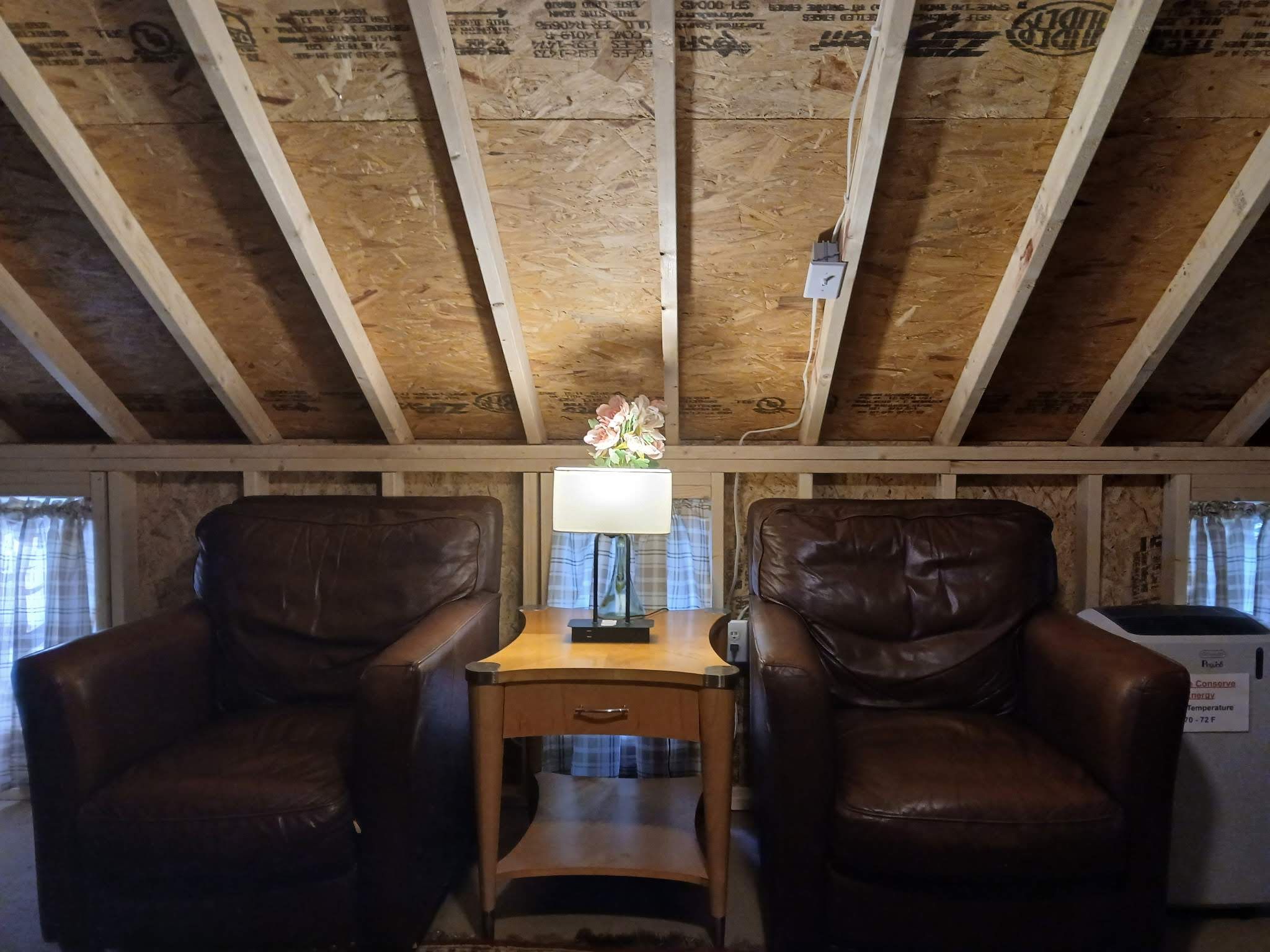A room with a wooden ceiling, two leather chairs, a small wooden table with a lamp, and a thermostat on the wall.