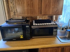 Two microwaves on a wooden countertop with a sink and a window in the background.
