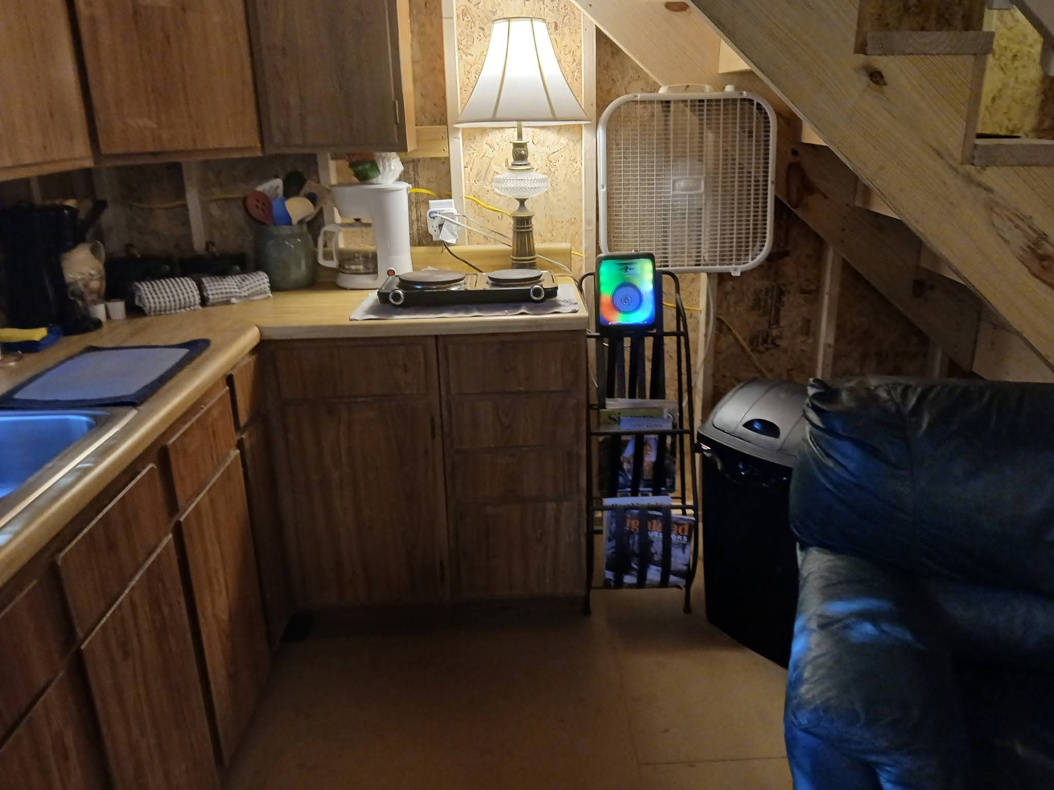 A compact kitchen with wooden cabinets, a small countertop, a coffee maker, a lamp, a fan, and a trash bin.