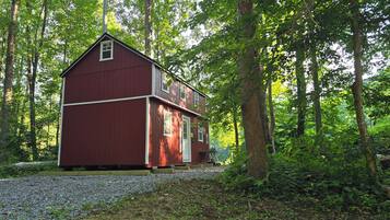 Une cabane en bois rouge, dotée d’un toit à pignon, entourée d’une forêt dense et bordée d’un chemin de gravier.