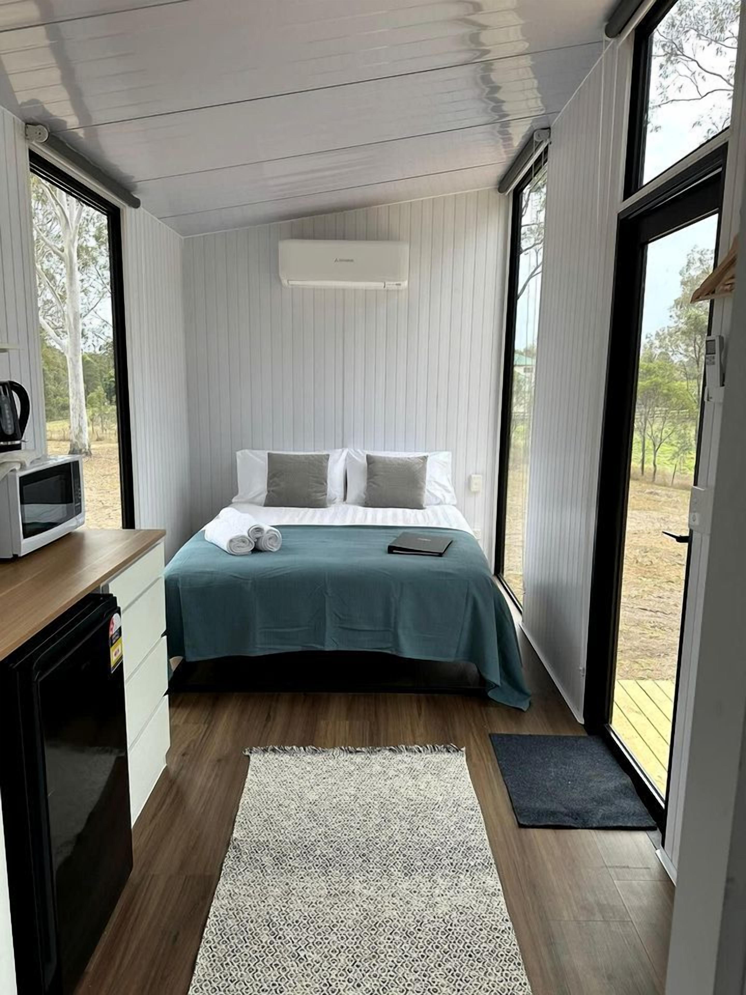 A bedroom with a bed, pillows, a bedside table with a microwave, and a view of the outdoors.