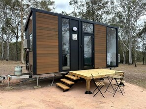 A modern, compact cabin with a wooden exterior and large windows, featuring a dining area with benches and a table.