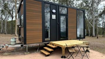 A modern, compact cabin with a wooden exterior and large windows, featuring a dining area with benches and a table.