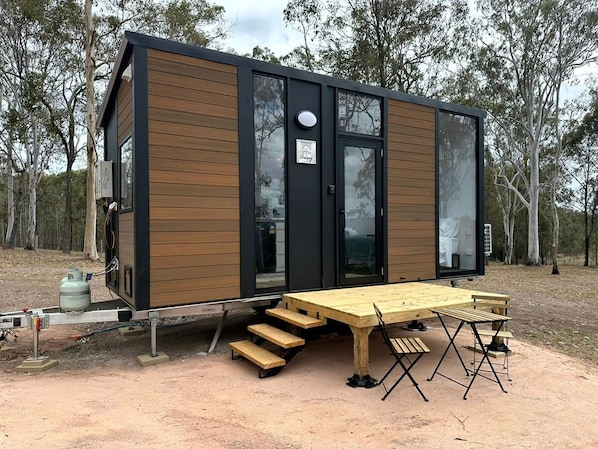 Una cabaña moderna y compacta con fachada de madera y grandes ventanales, que cuenta con una zona de comedor con bancos y mesa.