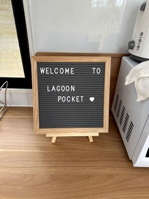 A welcome sign in a wooden frame on a wooden surface, next to a white appliance.