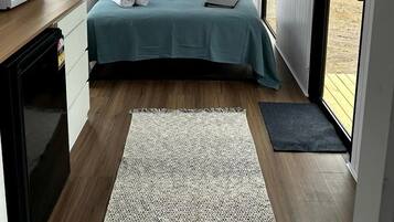 A bedroom with a bed, pillows, a bedside table with a microwave, and a view of the outdoors.