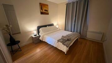 A bedroom with a bed, bedside tables, a nightstand, a radiator, a window with curtains, and a painting on the wall.