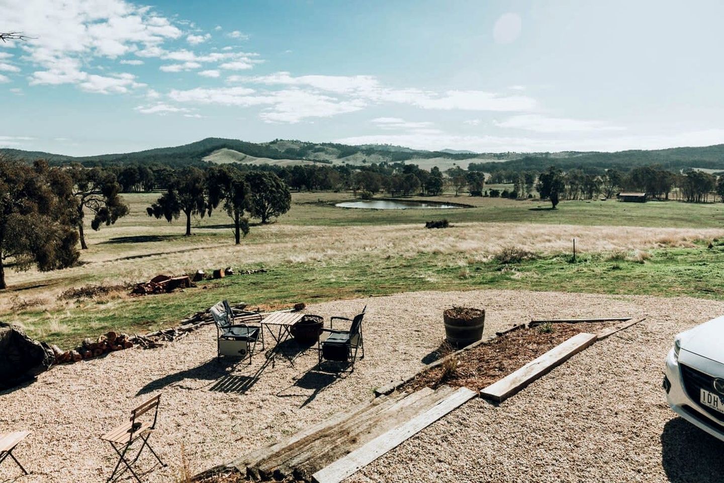 A gravel parking area with a white vehicle, outdoor seating, and a view of a hilly landscape.