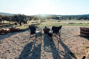 An outdoor seating area with two chairs and a table, surrounded by a gravel ground, trees, and a distant view of hills.