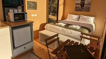 A compact room with a bed, a desk, and a chair. There's a window with blinds, a wall-mounted air conditioner, and a framed picture above the bed.