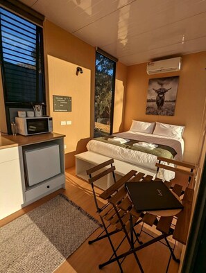 A compact room with a bed, a desk, and a chair. There's a window with blinds, a wall-mounted air conditioner, and a framed picture above the bed.
