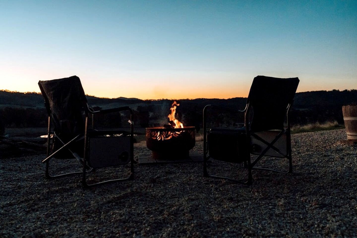 Two chairs facing a campfire with a view of a sunset over a hilly landscape.