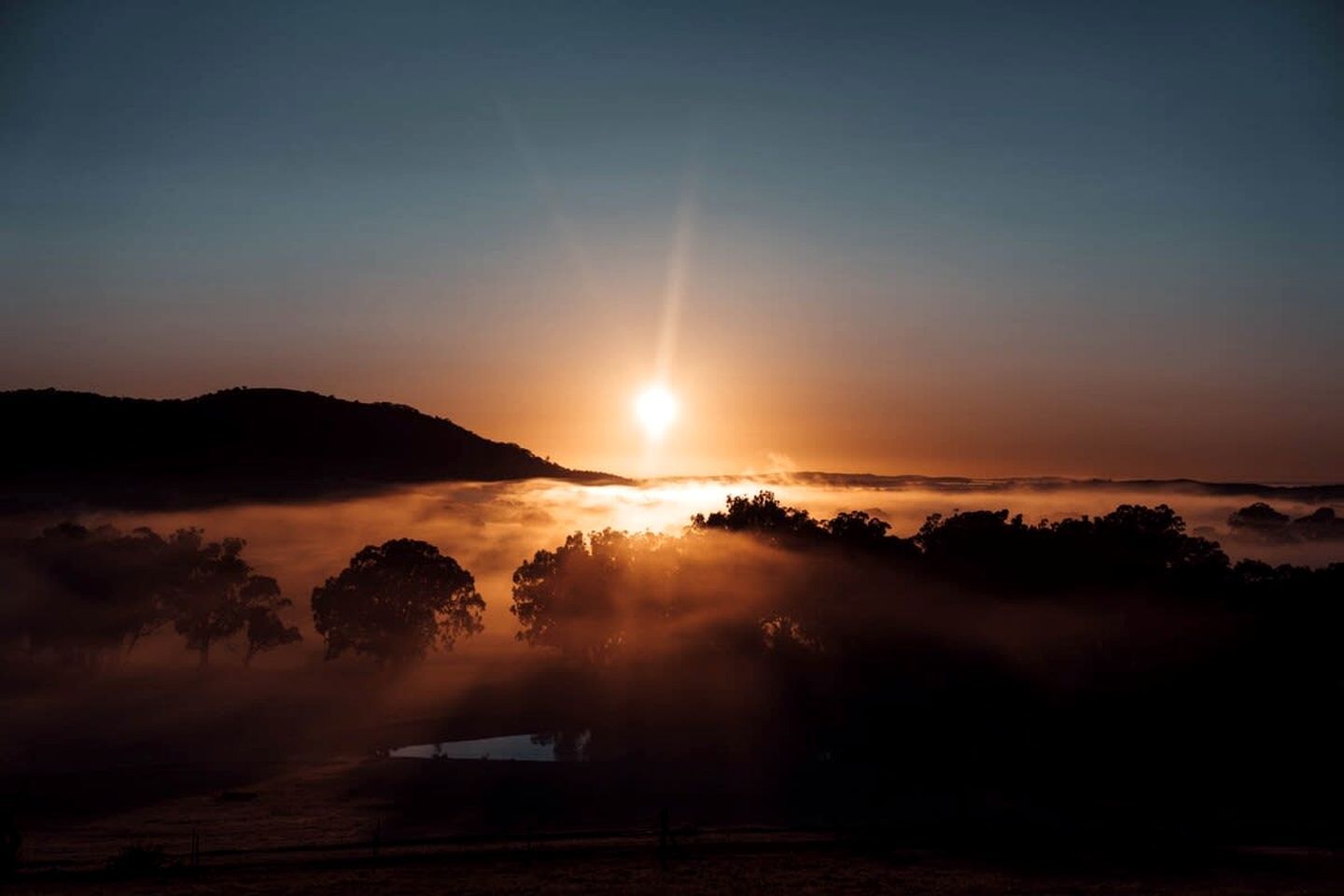 Een zonsopgang boven een mistig landschap met bomen en een heuvel in de verte.