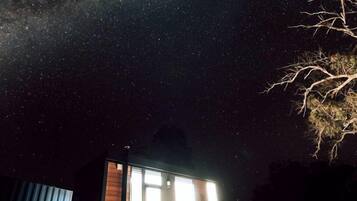 Una cabaña pequeña con ventana, una terraza de madera y un cielo nocturno estrellado.