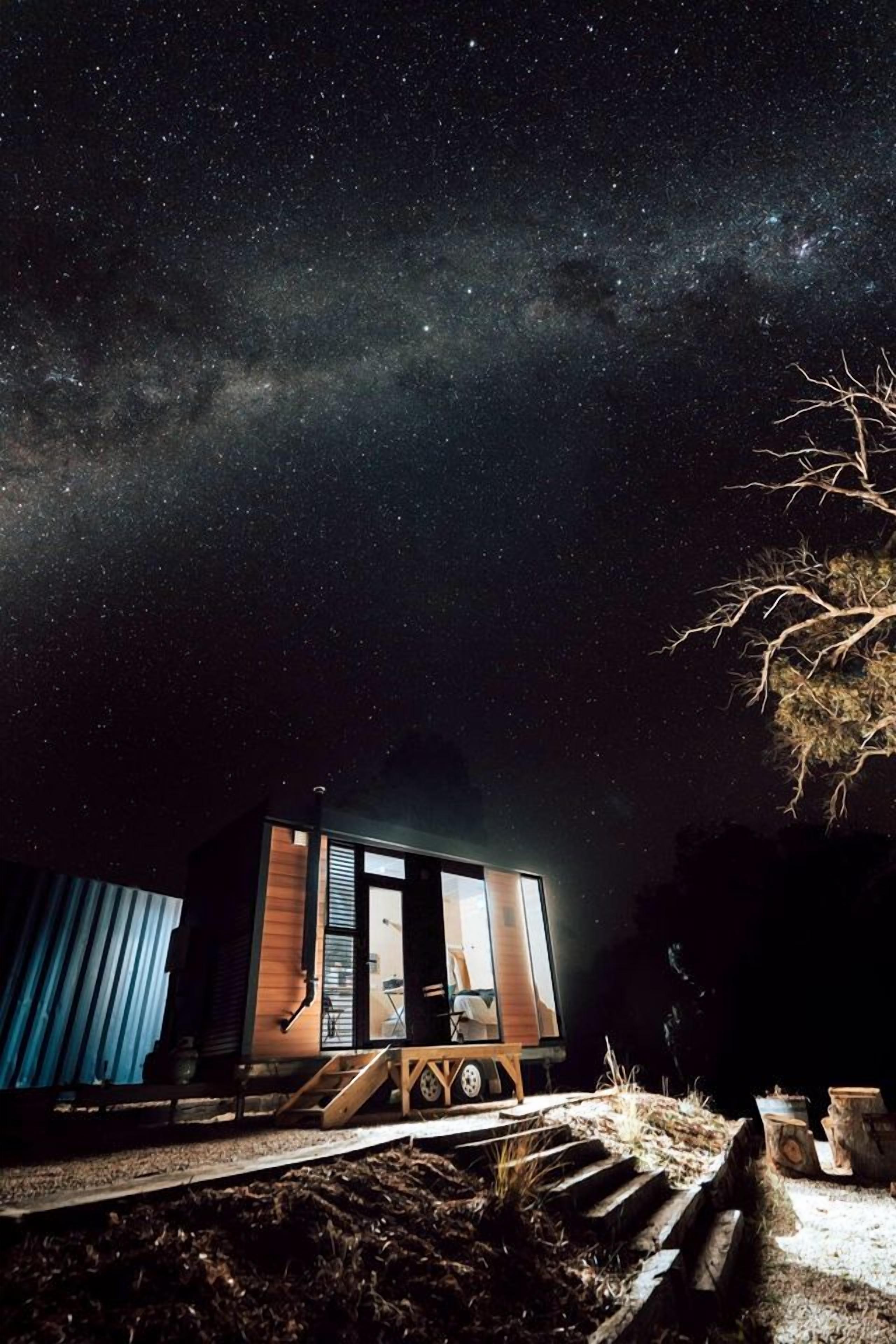A small cabin with a glass door, situated on a dirt path with a starry night sky above.