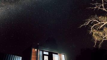 A small cabin with a glass door, situated on a dirt path with a starry night sky above.