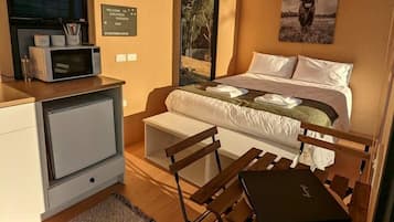 Habitación compacta con cama, escritorio y silla. Cuenta con ventana con persianas, aire acondicionado de pared y un cuadro enmarcado sobre la cama.