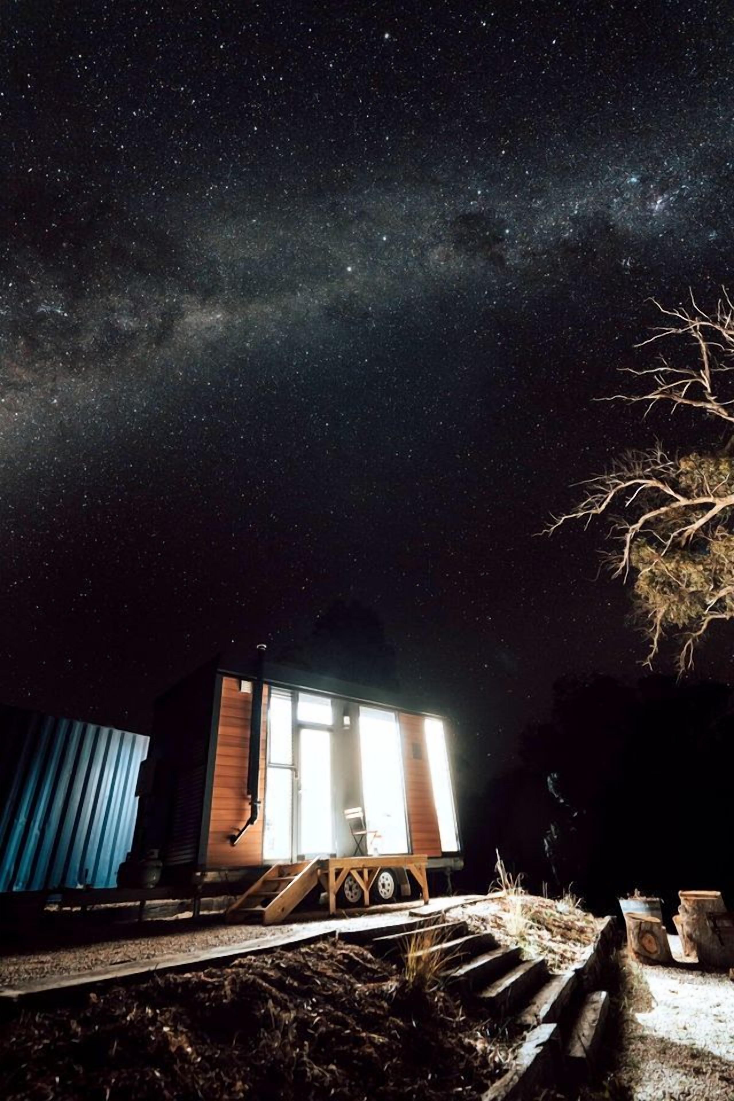 Una cabaña pequeña con ventana, una terraza de madera y un cielo nocturno estrellado.