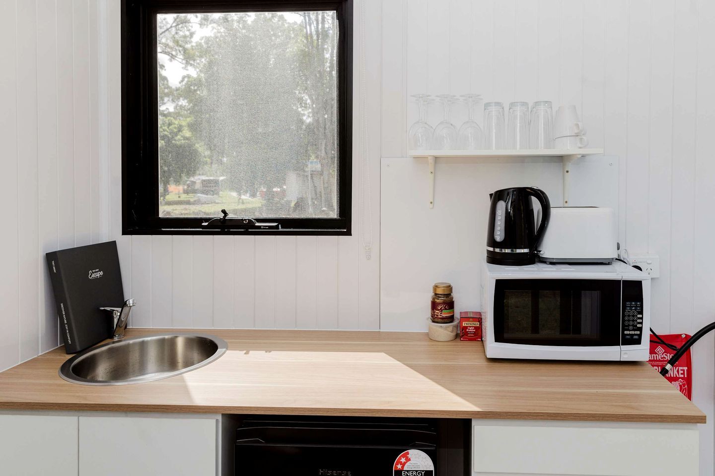Cocina compacta con un lavamanos de acero inoxidable, microondas, cafetera y ventana con vistas a árboles.