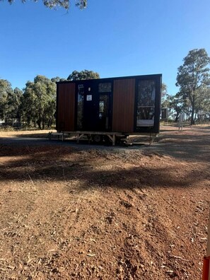 A small, single-story structure with a dark exterior and a visible door, situated on a dirt ground with sparse vegetation and trees in the background. - Cozy Tiny House Retreat Near Grampians National Park in Stawell Victoria (Stawell)