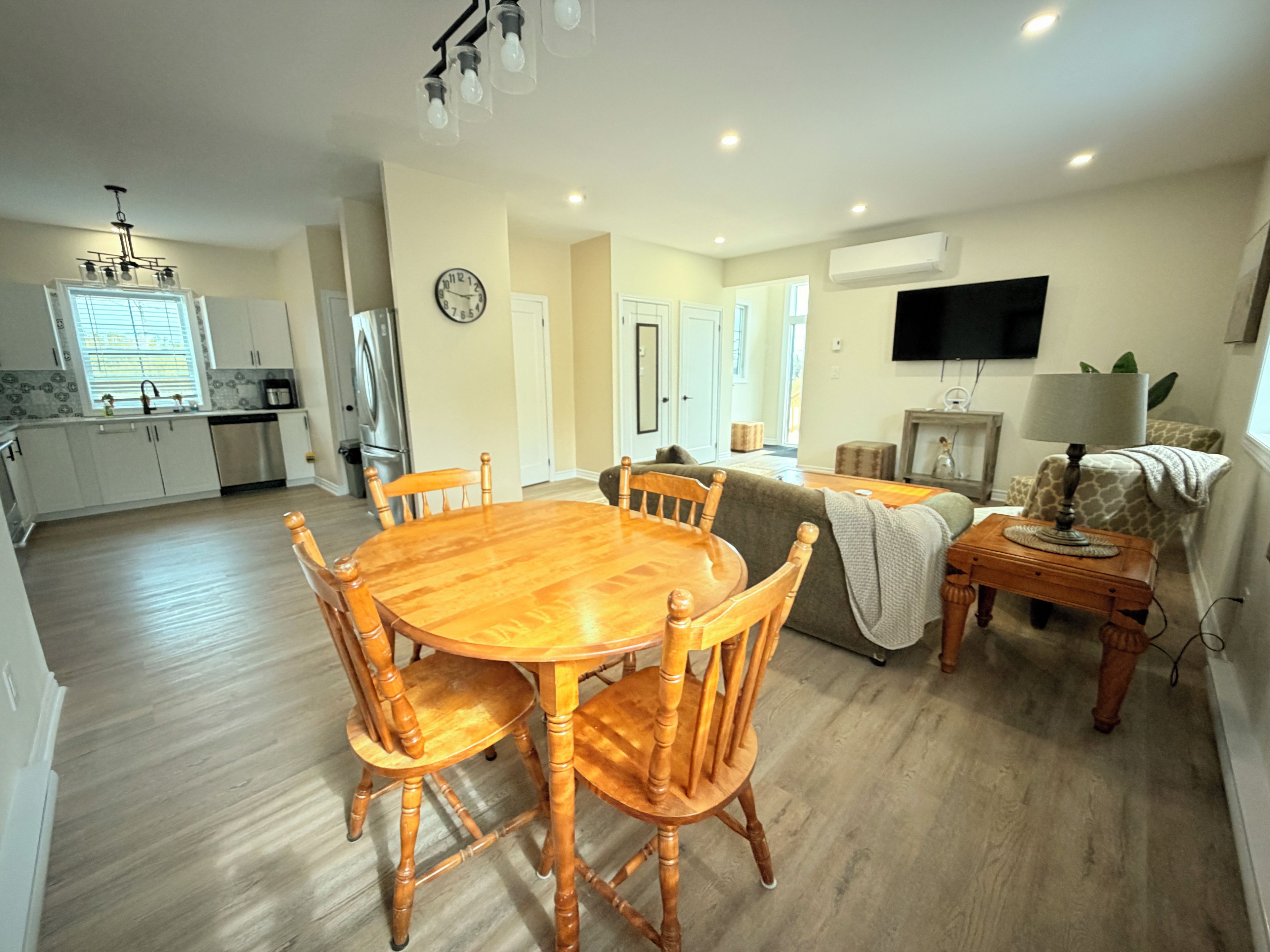 A modern kitchen with a dining area, a living room with a sofa and a television, and a hallway leading to bedrooms.