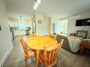 A dining area with a wooden table and chairs, a kitchen in the background, and a living room with a sofa and television.