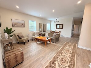 Un salón espacioso con una amplia mesa de centro de madera, un sillón estampado y un sofá. Hay dos ventanas con persianas, una zona de comedor con una mesa y sillas de madera, y un pasillo que conduce a otra habitación.