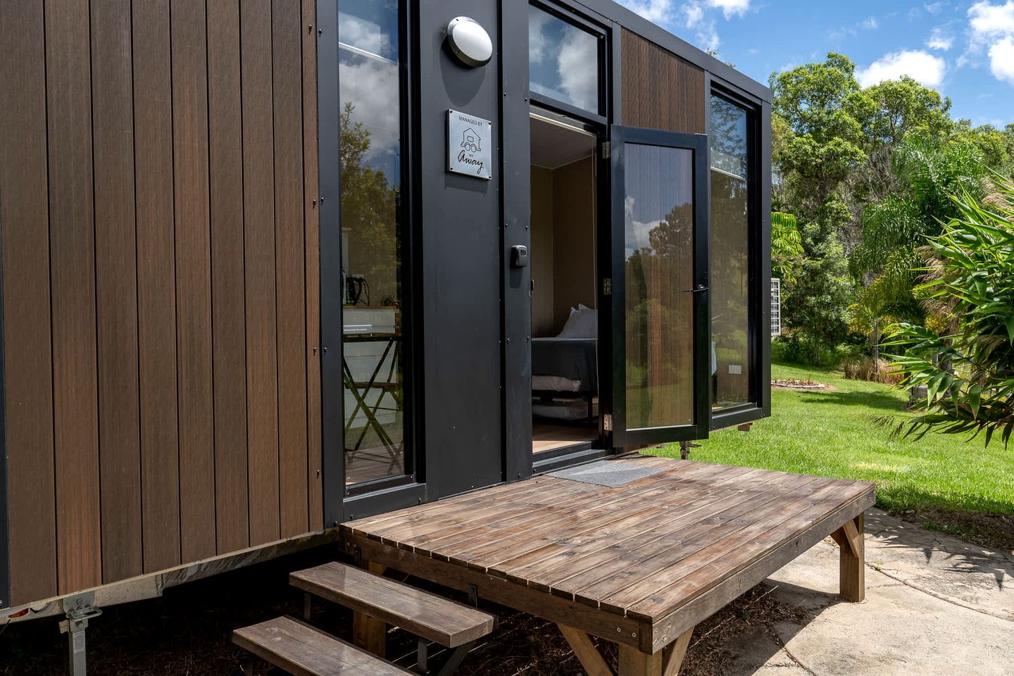 A modern, compact building with a wooden exterior, a large sliding glass door leading to a bedroom, and a wooden deck with a bench.