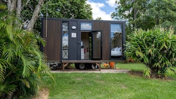 A modern, compact mobile home with a sleeping area and kitchenette, situated in a lush garden.