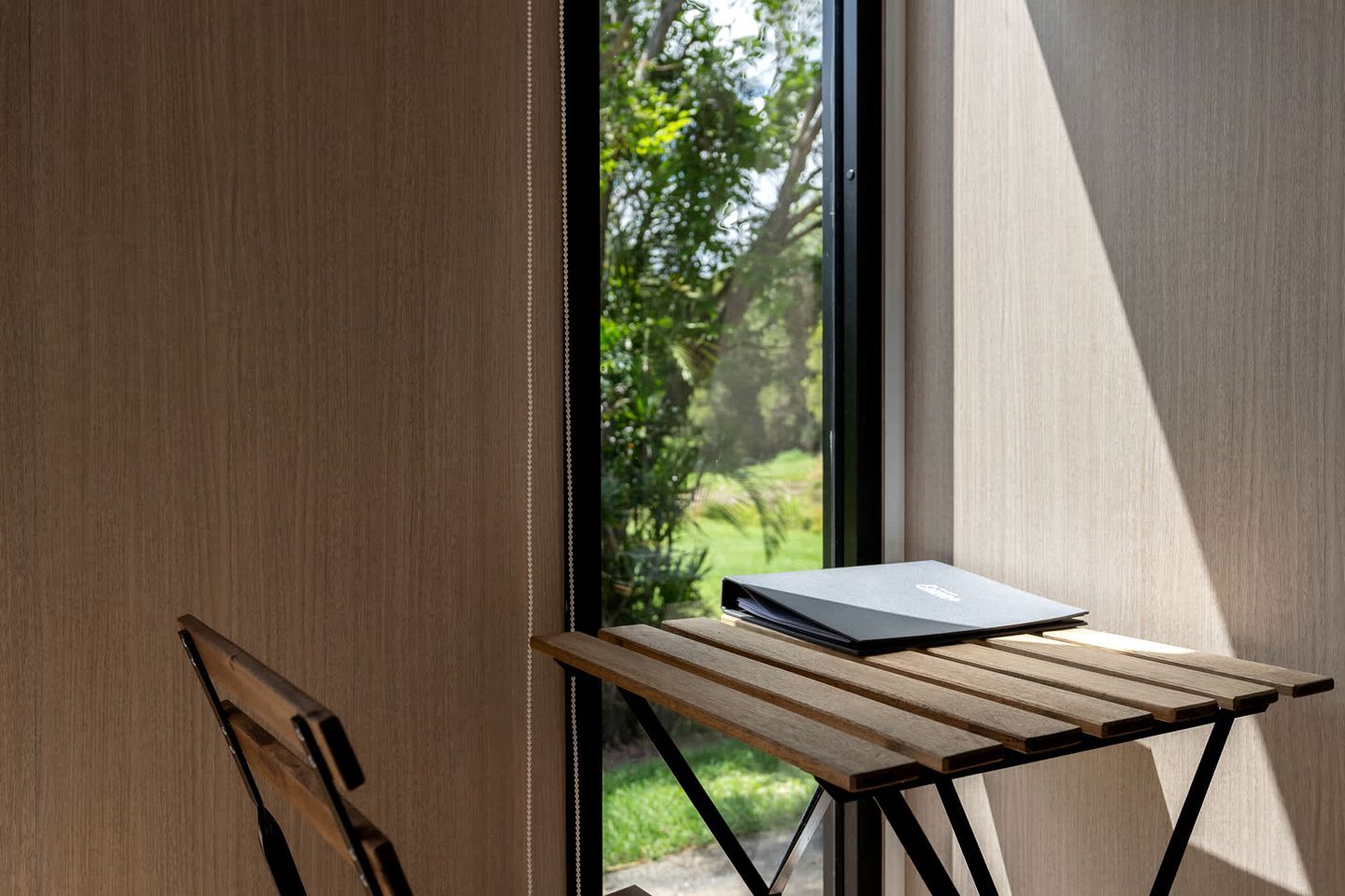 A wooden table with a laptop, a chair, and a view of greenery through a window.