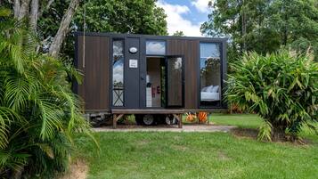 A modern, compact mobile home with a sleeping area and kitchenette, situated in a lush garden.