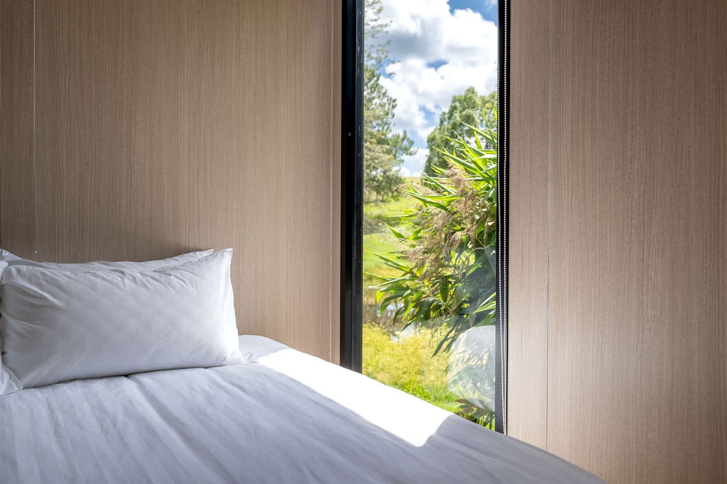 A bedroom with a large window, a bed with white bedding, and a view of greenery outside.