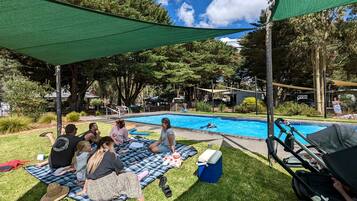 A group of people sitting on a blanket near a swimming pool, with a green shade sail overhead.