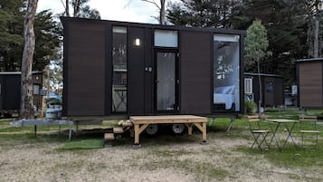 A compact, dark-colored mobile home with a sliding door, a small porch area, and a table with chairs.