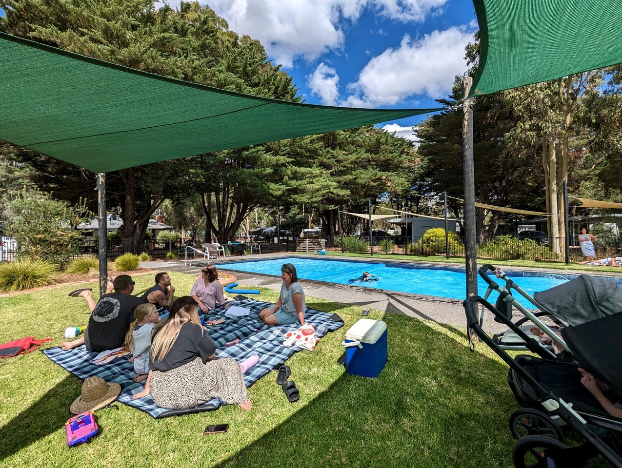 A group of people sitting on a blanket near a swimming pool, with a green shade sail overhead.