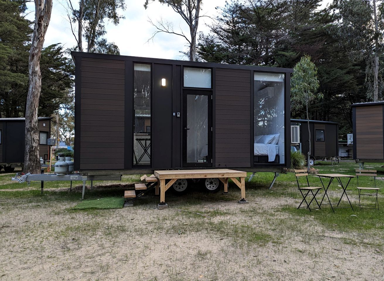 A compact, dark-colored mobile home with a sliding door, a small porch area, and a table with chairs.