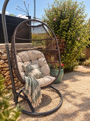 A hanging chair with a cushion and a throw blanket, set against a stone wall and surrounded by greenery.