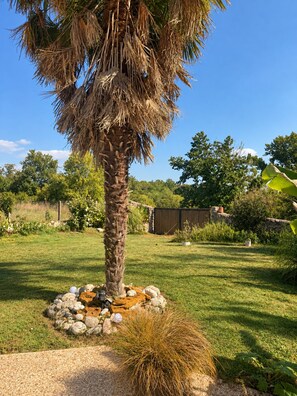 A palm tree with a thatched top surrounded by rocks and pebbles, set in a grassy yard with a stone wall and various plants in the background.