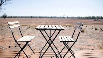 A table and two chairs set on a wooden deck with a dirt field and trees in the background.