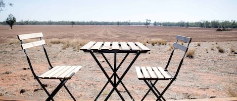 A table and two chairs set on a wooden deck with a dirt field and trees in the background.