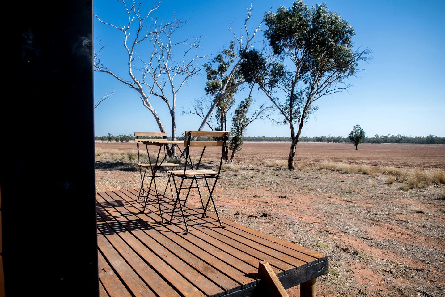 Un espace extérieur aménagé avec une terrasse en bois et des chaises en métal, offrant une vue sur un paysage aride et dégagé, parsemé de quelques arbres.