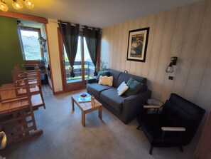 A living room with a sofa, a coffee table, and a wooden chair. There is a window with curtains, a picture on the wall, and a view of mountains outside. - Apartment in Les Coches with a View of Mont Blanc (La Plagne Tarentaise)