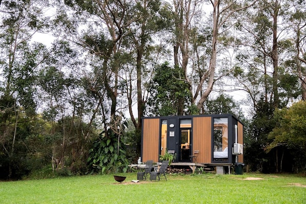 A modern, compact cabin with a wooden exterior and a black door, surrounded by lush greenery and trees.