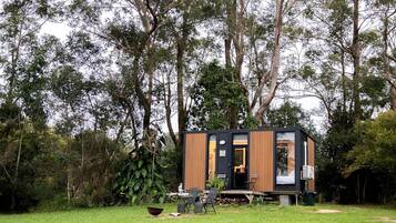 Une cabane moderne et compacte, dotée d’une façade en bois et d’une porte noire, entourée d’une végétation luxuriante et d’arbres.