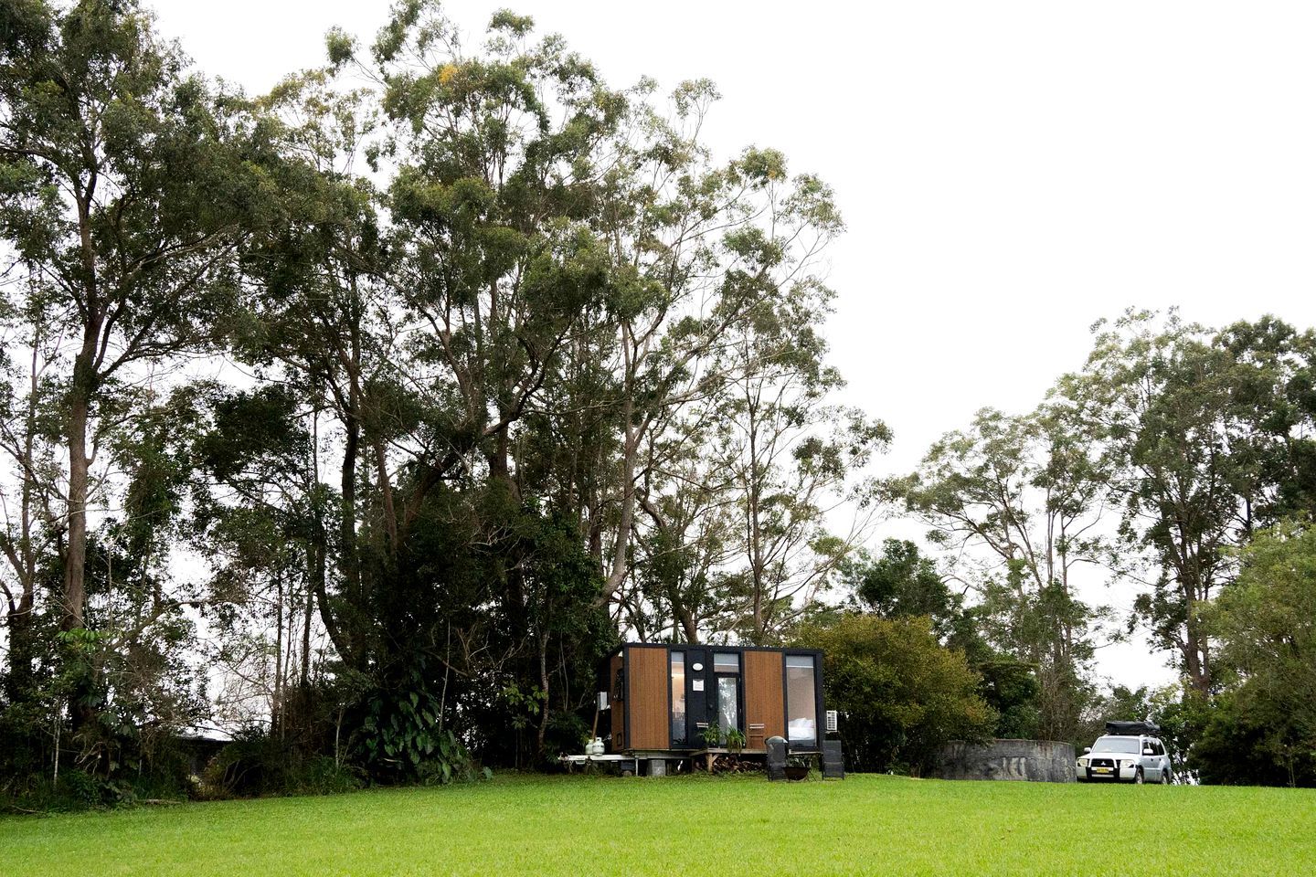Une cabane moderne et compacte, dotée d’une façade en bois, entourée d’une pelouse verdoyante et luxuriante et de grands arbres.
