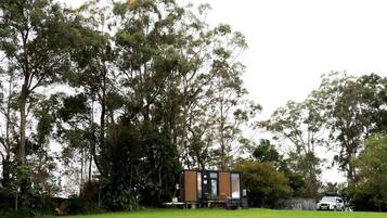 Une cabane moderne et compacte, dotée d’une façade en bois, entourée d’une pelouse verdoyante et luxuriante et de grands arbres.