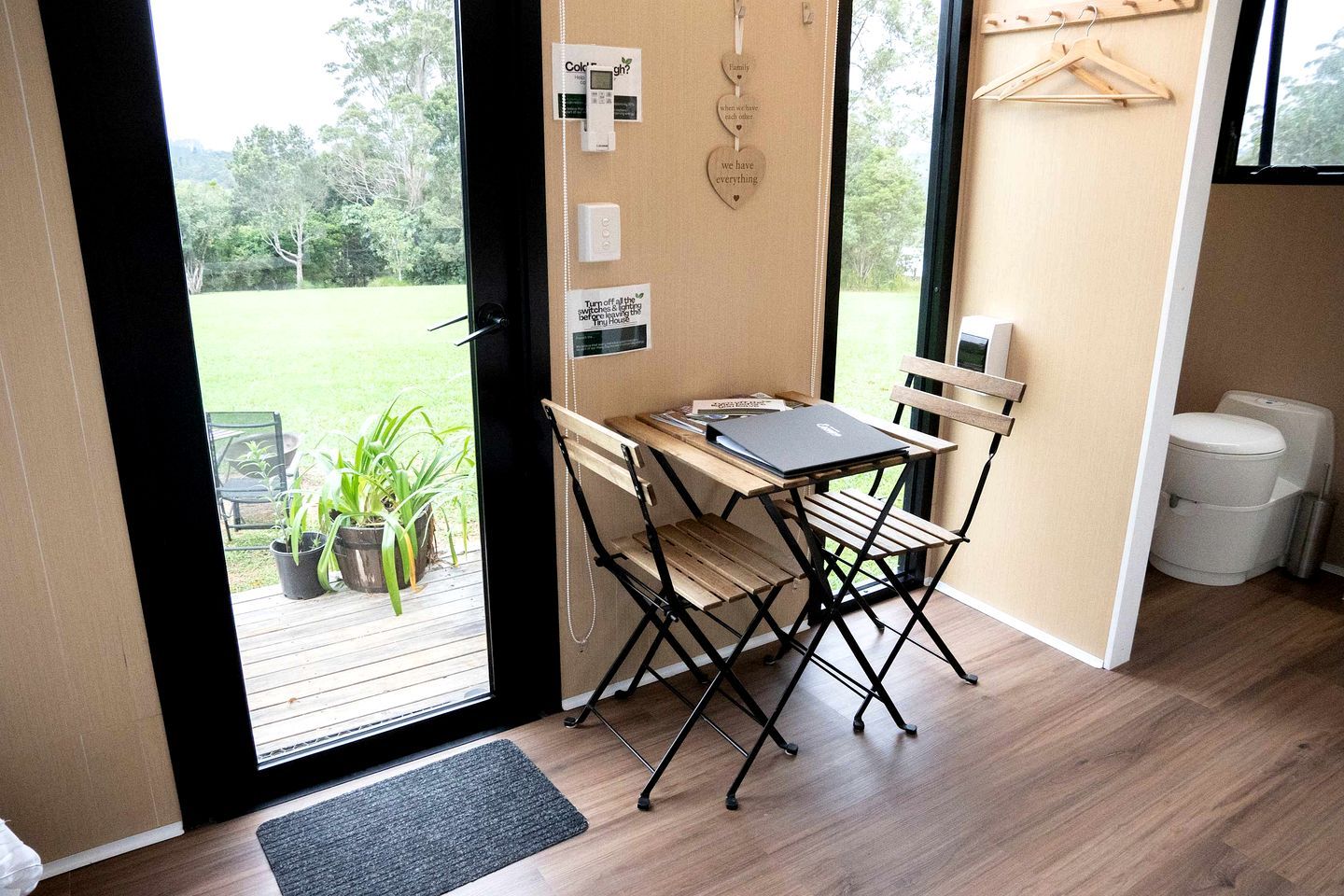 A room with a wooden desk and chairs, a laptop, a small potted plant, and a view of a green outdoor area.