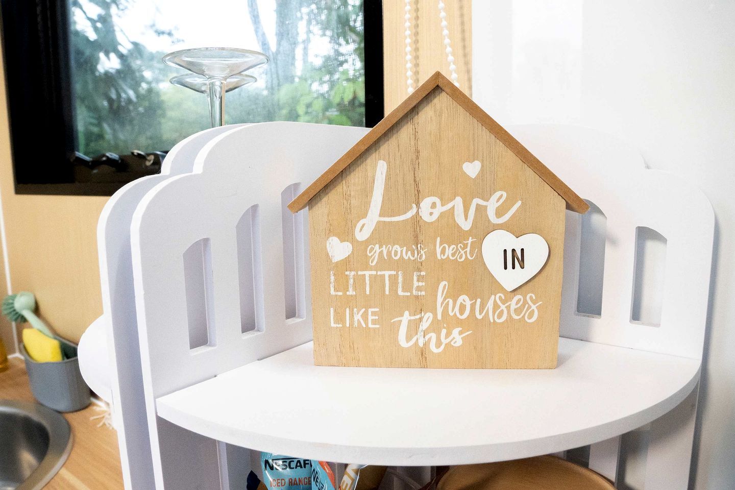 Une enseigne en bois en forme de maison, sur laquelle on peut lire : « L’amour s’épanouit le mieux dans les petites maisons comme celle-ci », et qui est posée sur une chaise blanche.