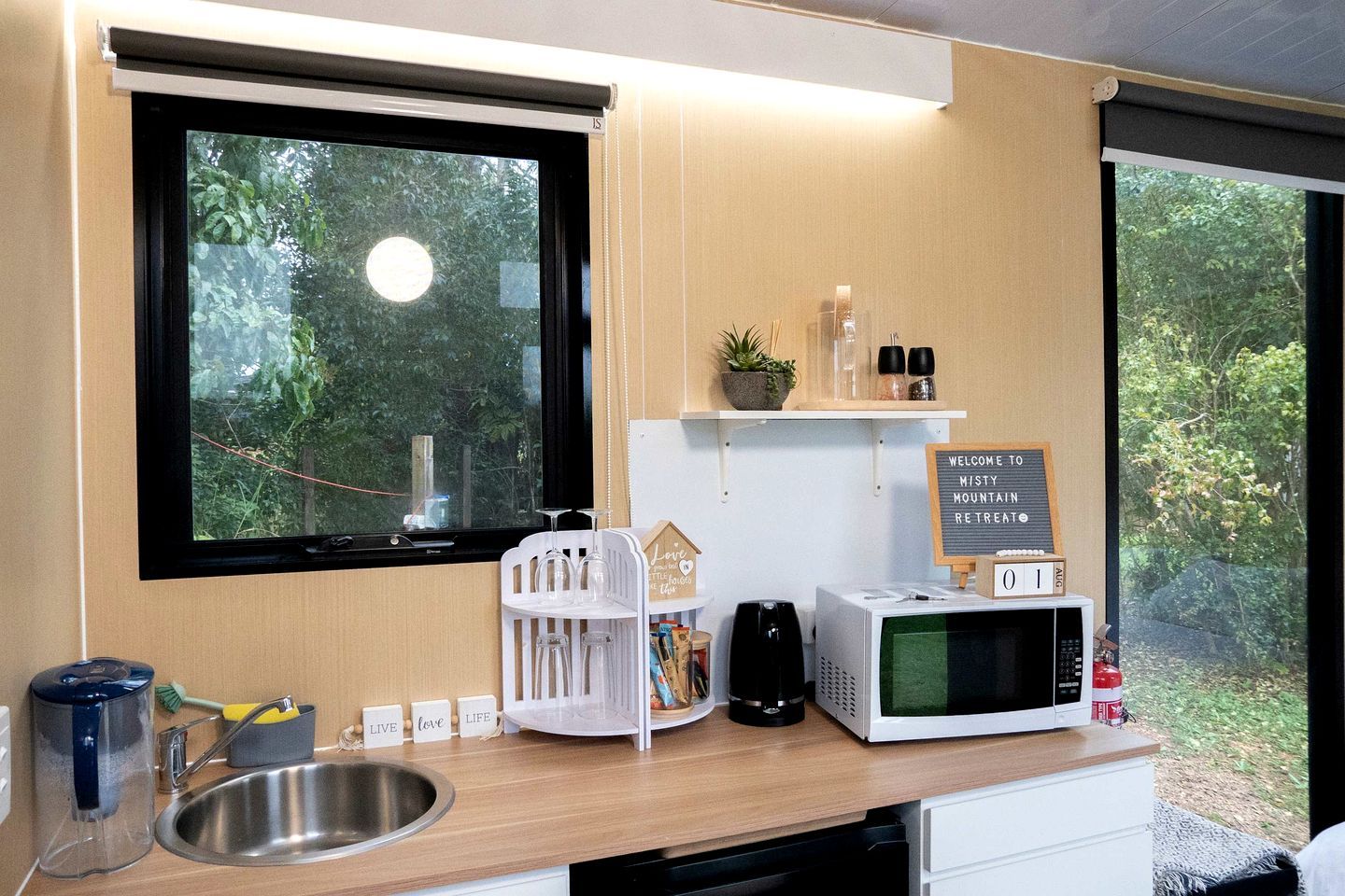 A kitchen with a sink, a microwave, a coffee maker, and a sign that reads 'Welcome to Misty Mountain Retreat.'