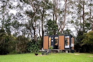 Una cabana moderna i compacta, amb façana de fusta i porta negra, envoltada de verdura abundant i arbres.